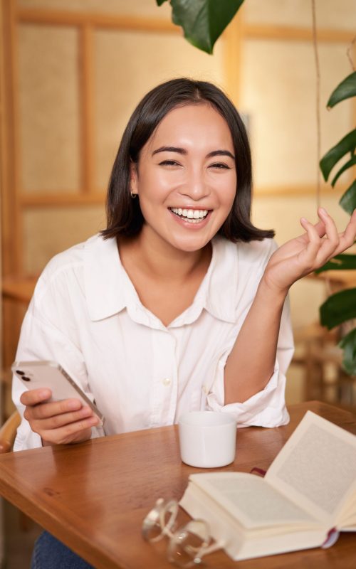 Stylish young woman in cafe with coffee, holding mobile phone, laughing and smiling. Lifestyle and people concept.